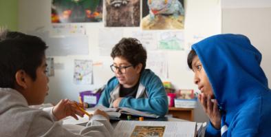 three kids sitting at desks talking and working