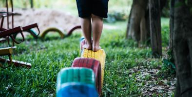 Child standing on a tire that is painted yellow. The yellow tire is 4th in line after a blue, green and red tire.
